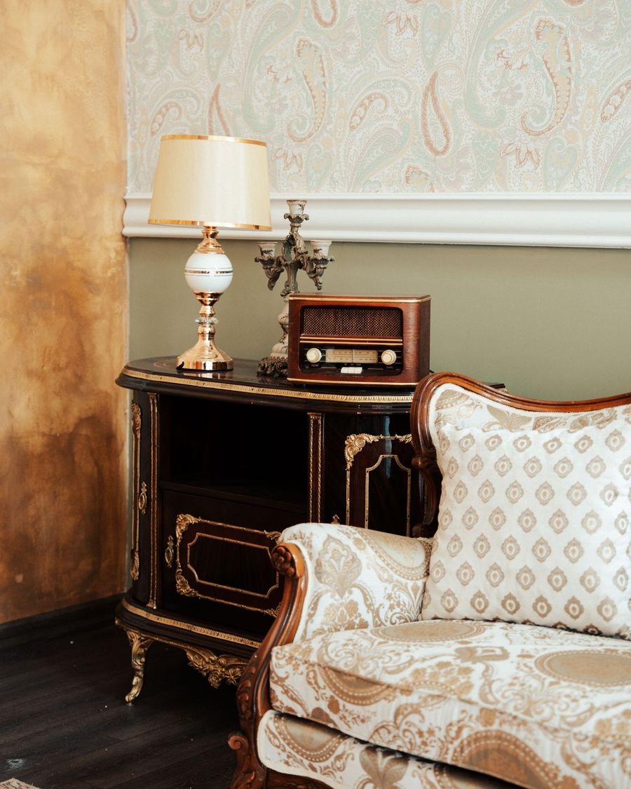 Sitting corner with a soft paisley wallpaper above a white chair rail, an upholstered settee, an antique radio, and a brass table lamp