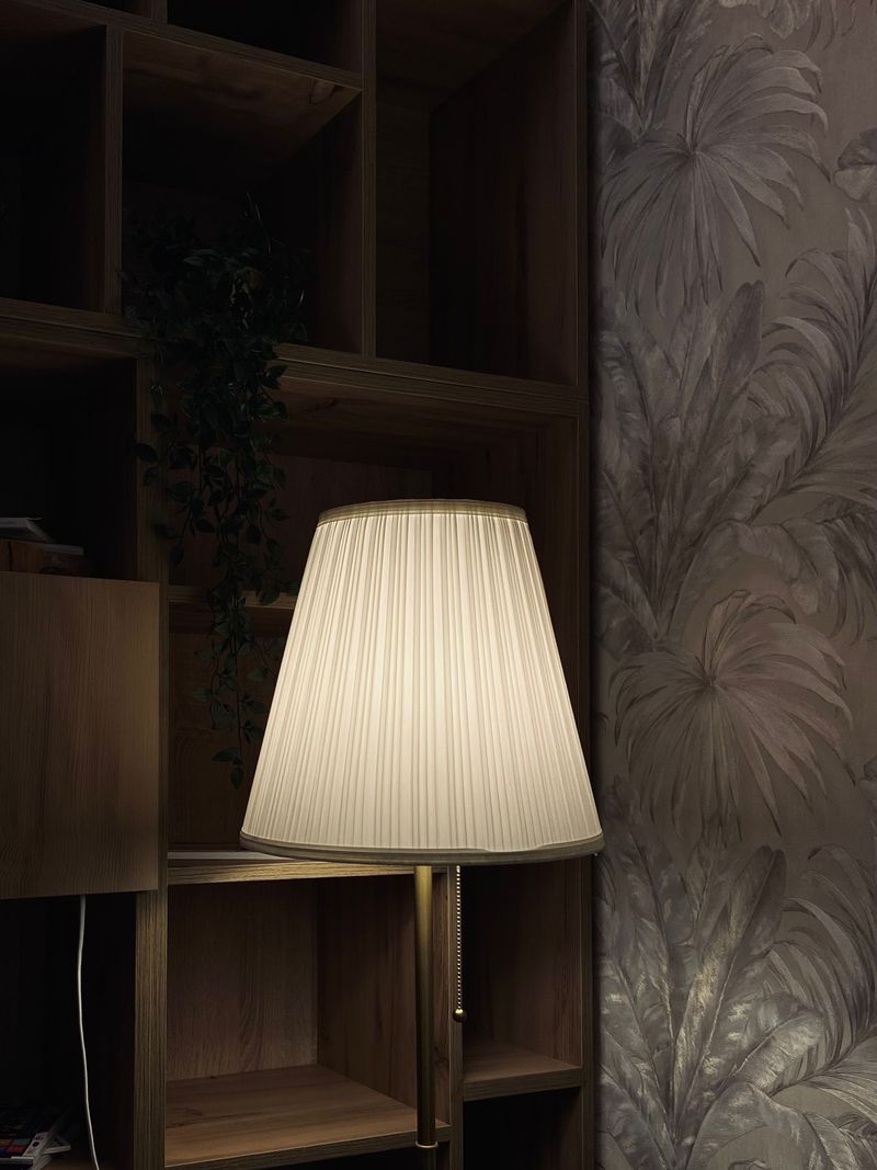 Pleated floor lamp glowing beside dark wood shelving and a gray embossed leaf wallpaper