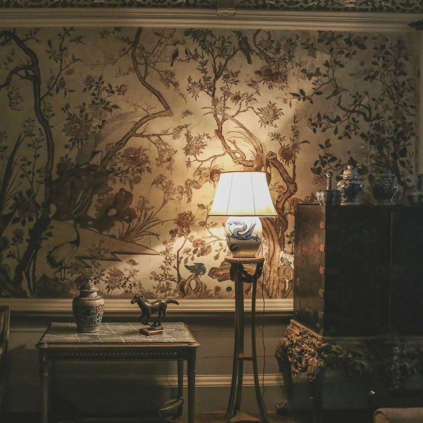 A living room wrapped in a hand-painted chinoiserie mural wallpaper of flowering branches, softly lit by a single table lamp beside antique furniture