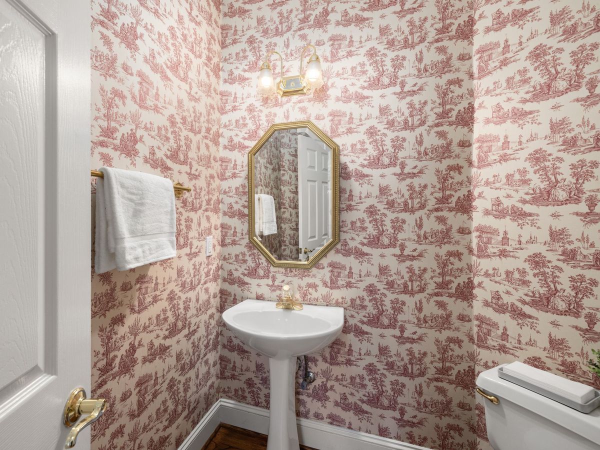 View through a doorway into a powder room papered in red toile, with a pedestal sink and an octagonal gold mirror
