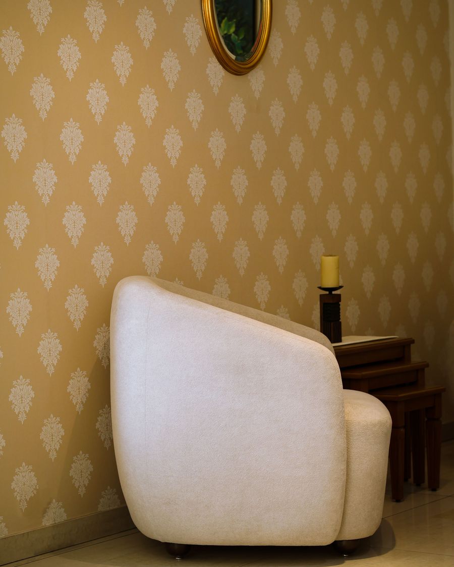Quiet corner with a golden damask wallpaper, a white tub armchair, a small side table with a candle, and an oval mirror