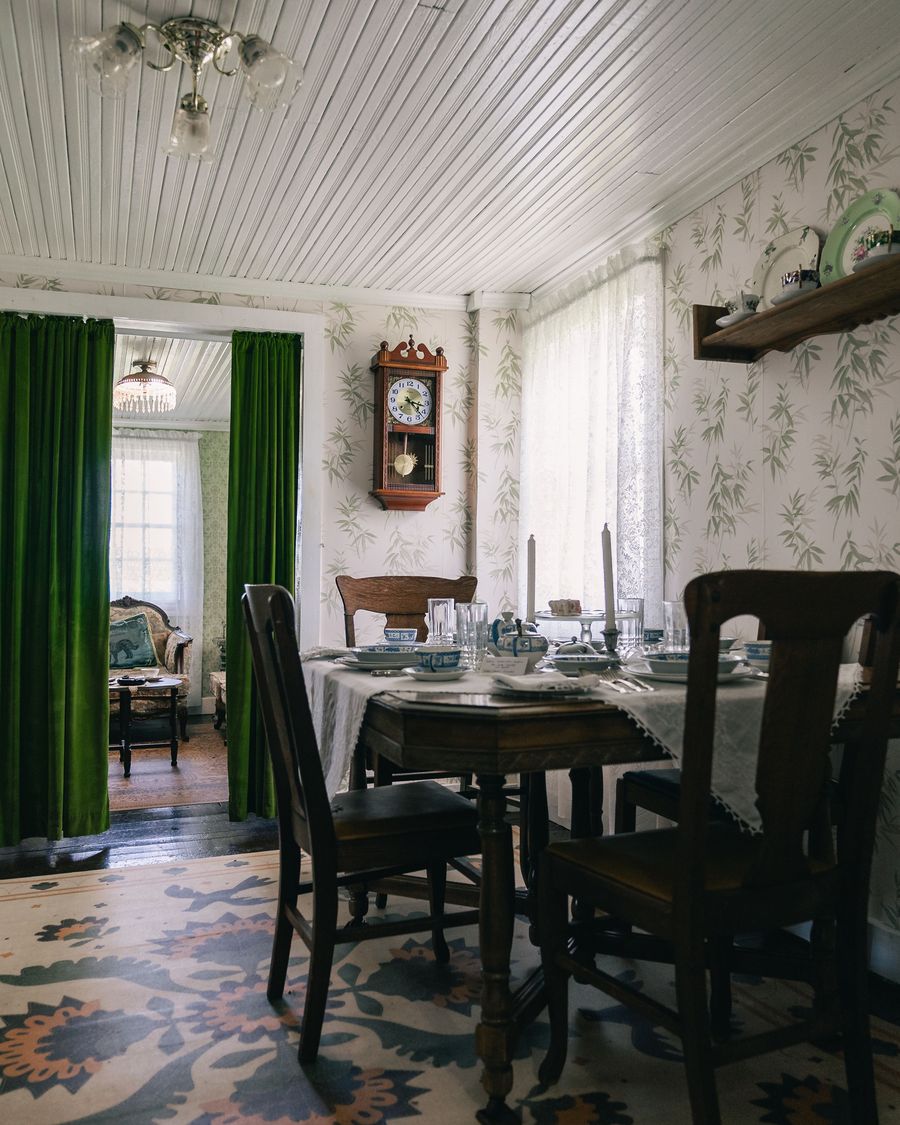 Traditional dining room with pale bamboo-leaf wallpaper, a set wooden table, a wall clock, and green velvet curtains