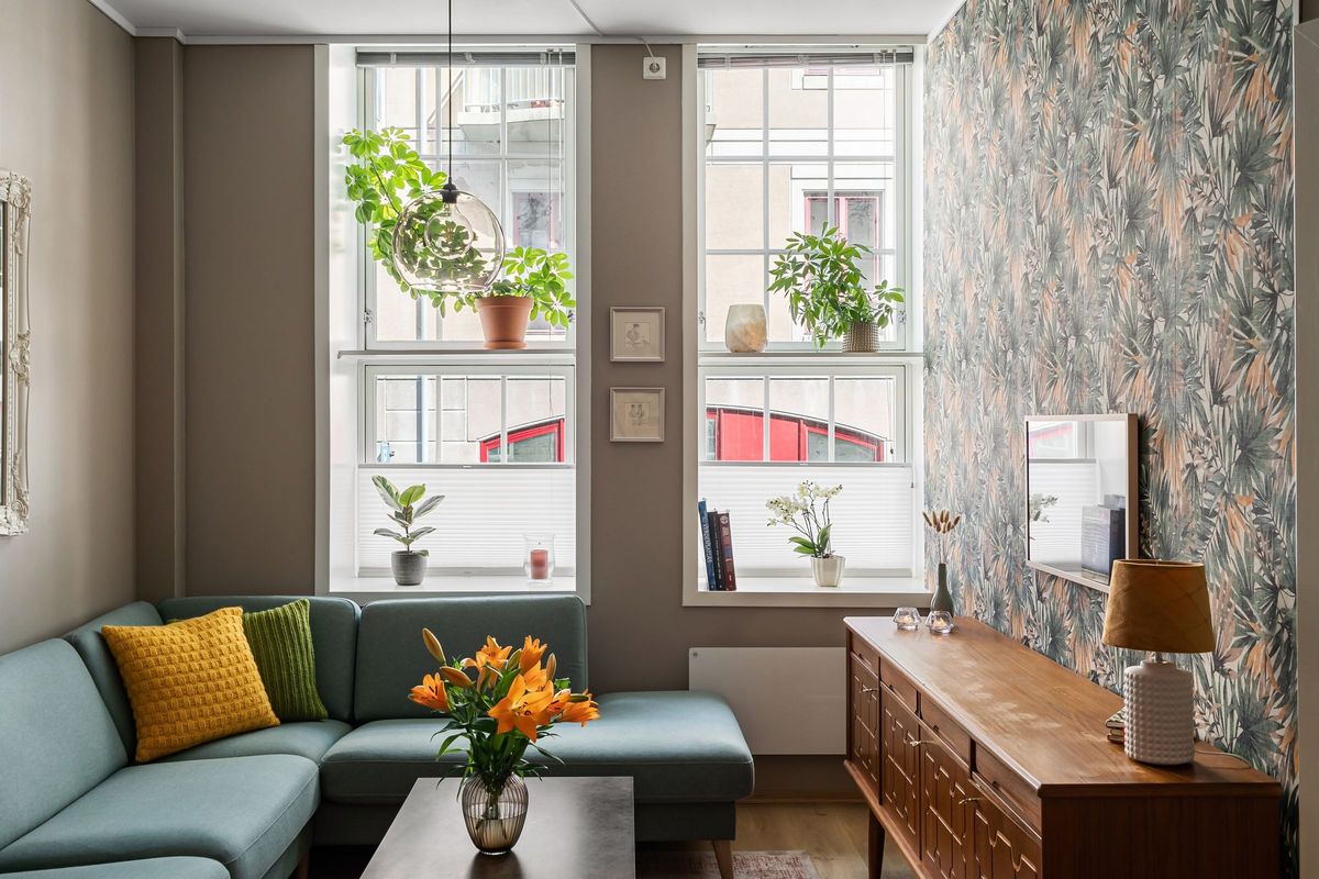 Bright living room with a botanical leaf wallpaper on the side wall, a teal sectional sofa, and tall windows lined with houseplants