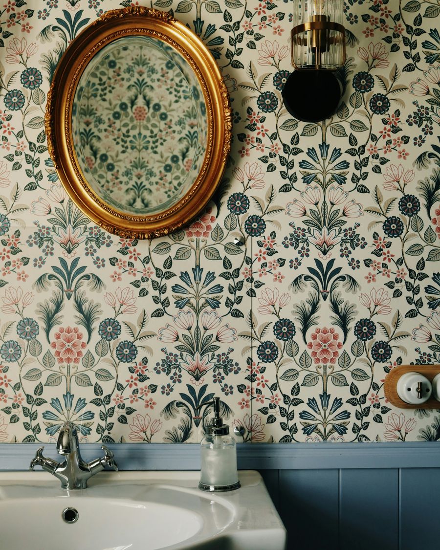 Bathroom with a cream folk-floral wallpaper above blue beadboard, an oval gilt mirror, a wall sconce, and a white sink