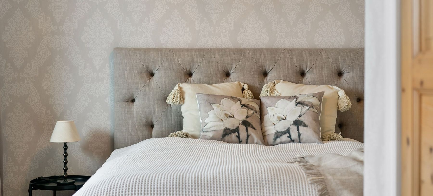 Calm bedroom with a soft beige damask wallpaper behind a tufted linen headboard, white bedding, and a small bedside lamp